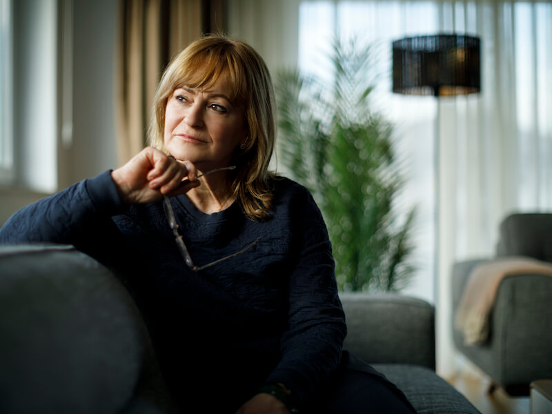 Woman sitting on a grey couch gazing out the window wondering if she has hearing loss.