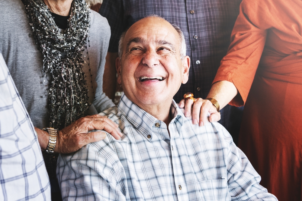Elderly man smiling, surrounded by group of adults touching his shoulder.