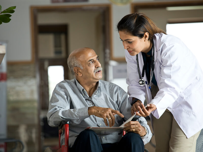 Female doctor communicating with older man who has hearing loss in wheelchair examining reports at the hospital corridor.